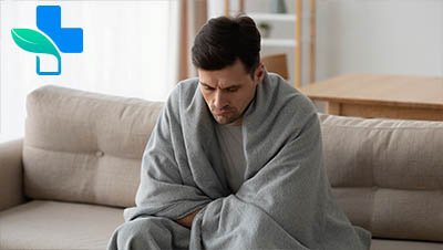 Мужчина сидит на диване, укутанный в одеяло, и грустно смотрит вниз
