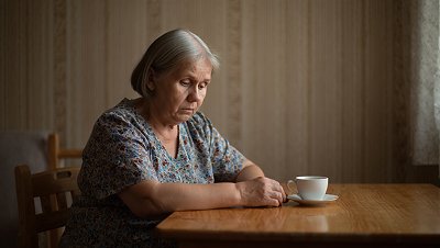 Пожилая женщина сидит за столом и грустит